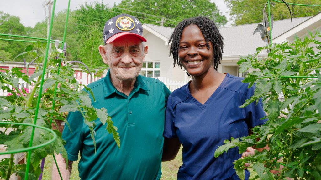 A nurse and resident at a skilled nursing home smile at the camera surrounded by a garden